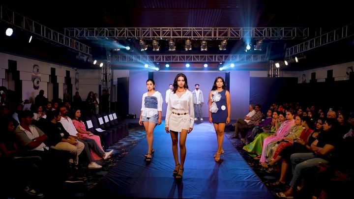 Three finalists walk the runway under blue light, audience on both sides.