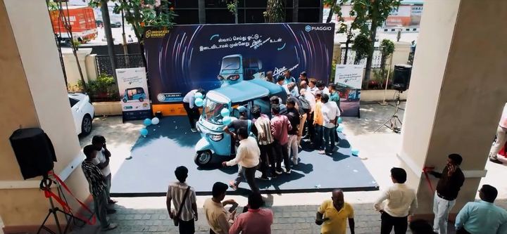 A crowd gathers around the unveiled three-wheeler, seen from above.