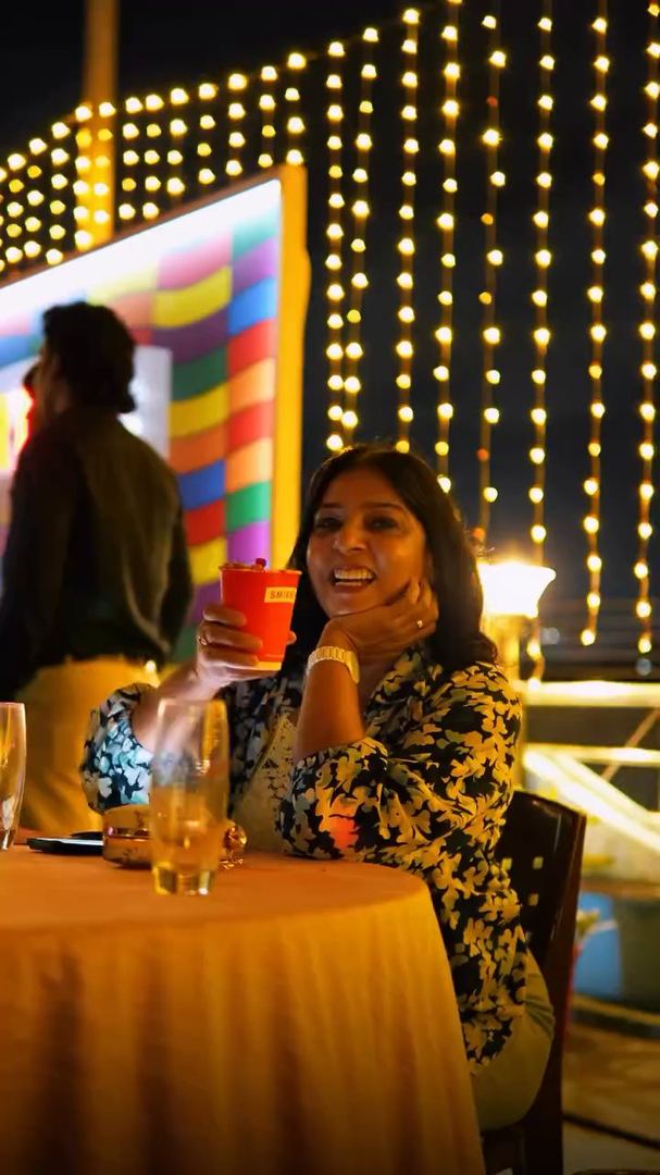 Guest smiles at her table with a drink under string lights.